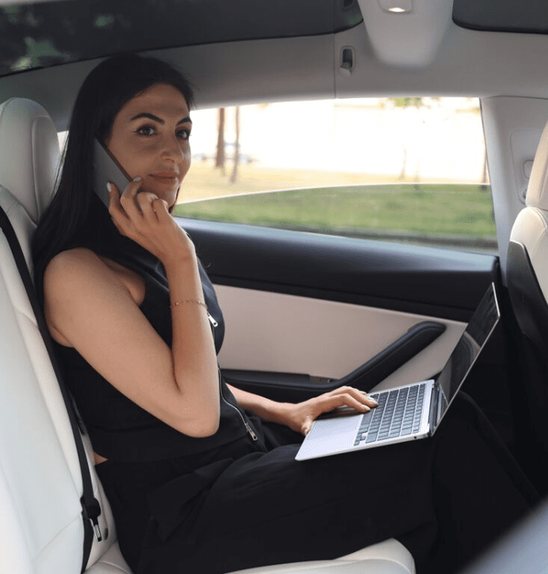 Woman using laptop and phone in car.